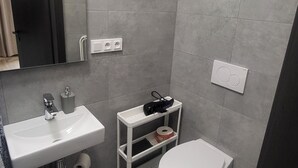 Ein modernes Badezimmer mit einem weißen Waschbecken, einem Spiegel und einer Toilette. Eine kleine weiße Ablagefläche mit einer schwarzen Tasche und Toilettenpapier.
