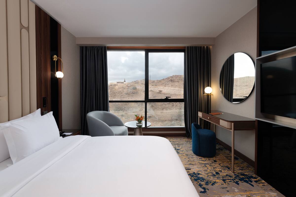A hotel room with a large bed, a desk, a chair, a TV, and a view of a desert landscape.