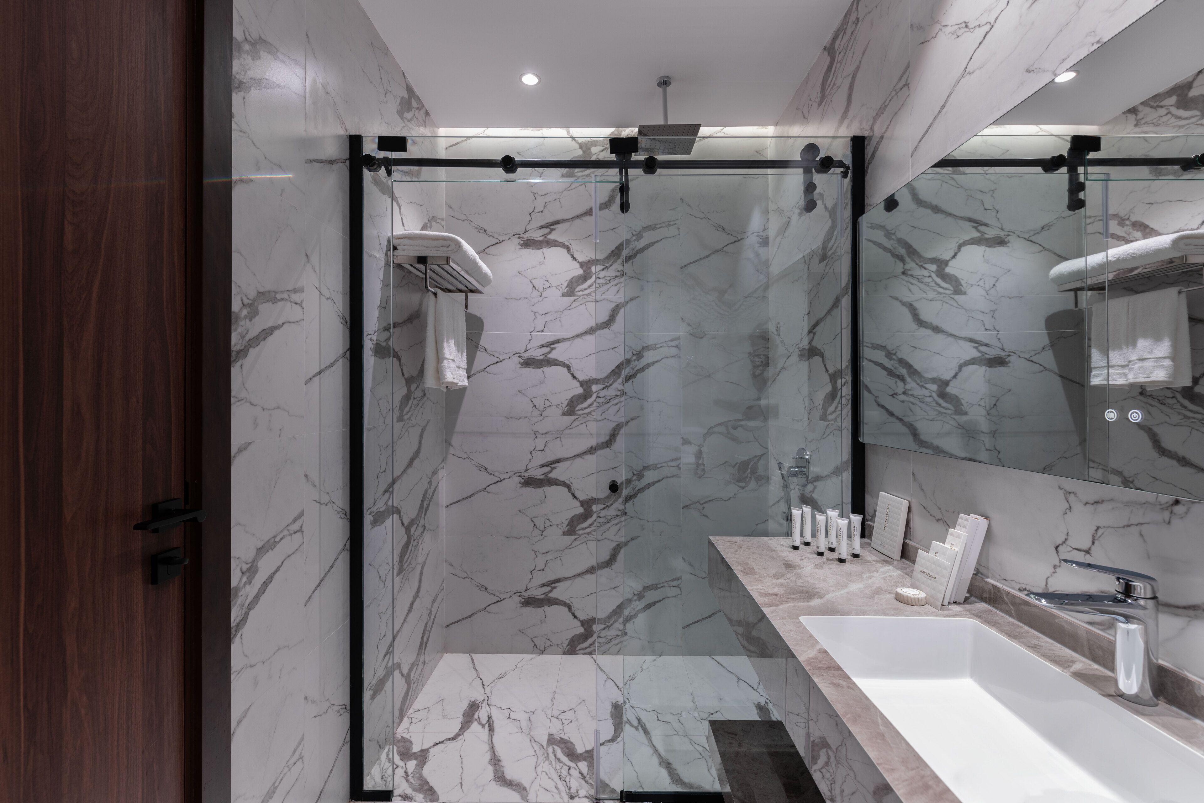 A modern bathroom with a marble wall, a glass shower enclosure, and a vanity with a sink and mirror.