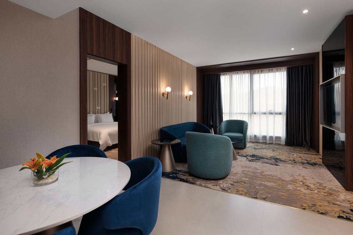 A modern hotel room with a round dining table, blue chairs, a small side table, a large window with curtains, and a view of the city.