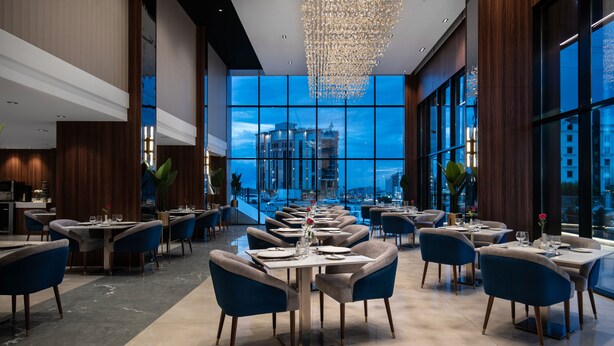 A modern restaurant with a view of a cityscape through large windows. Tables are set with white tablecloths and blue chairs.