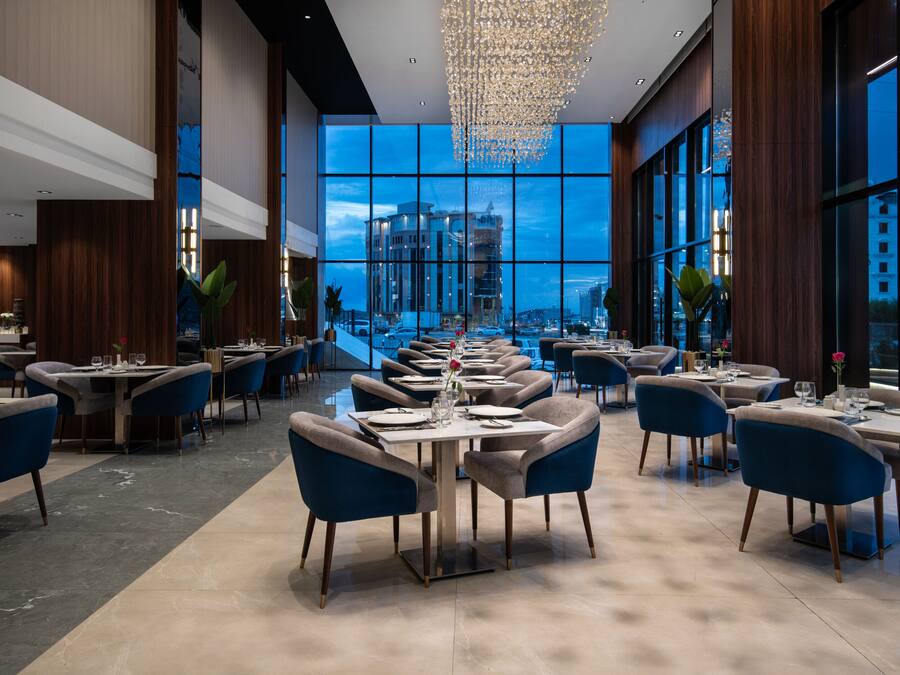 A modern restaurant with a view of a cityscape through large windows. Tables are set with white tablecloths and blue chairs.