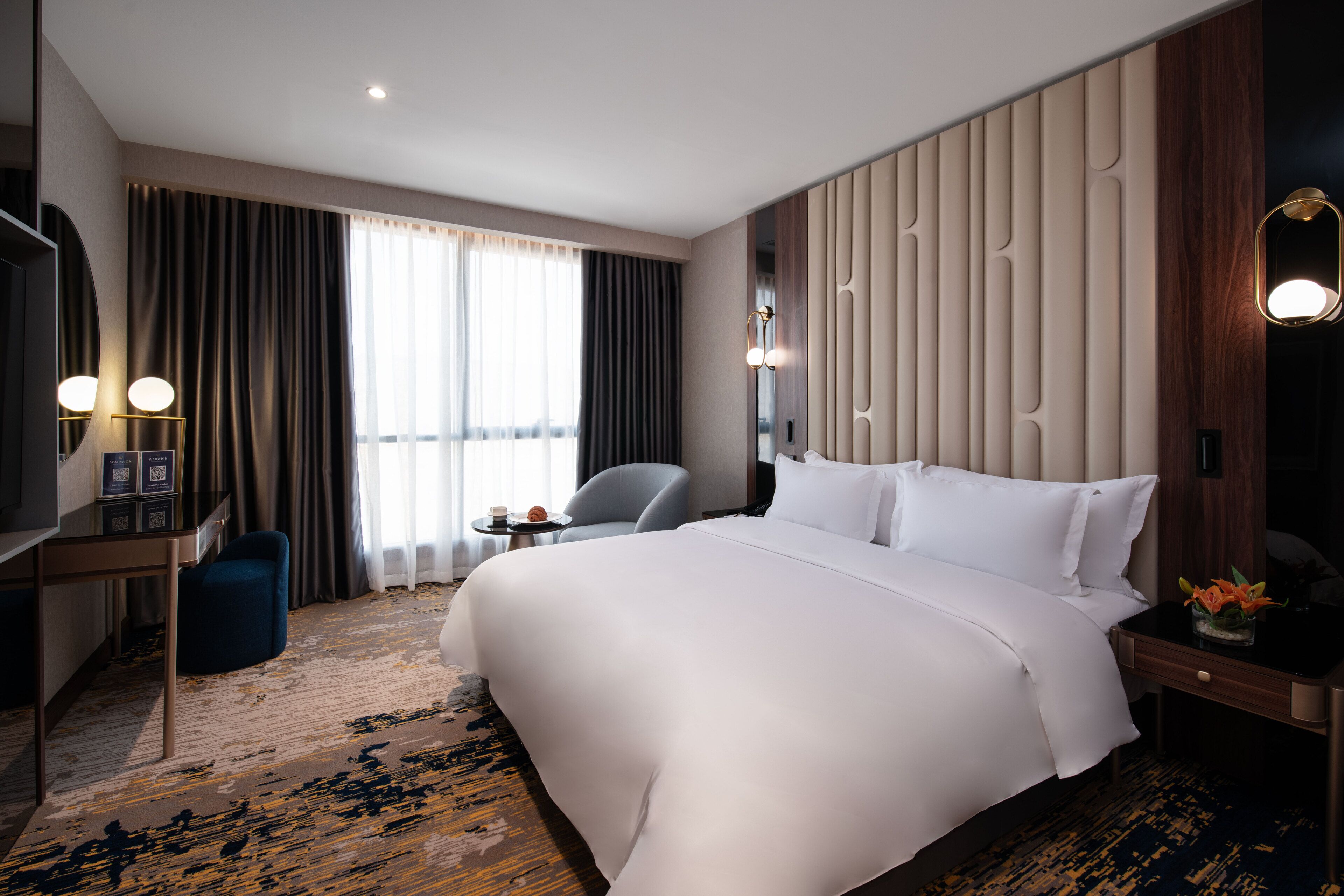 A hotel room with a large bed, a desk with a chair, a small table with a lamp, and a large window with curtains.