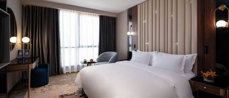 A hotel room with a large bed, a desk with a chair, a small table with a lamp, and a large window with curtains.