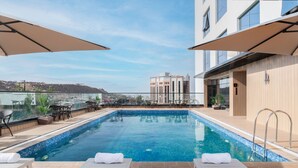 A rooftop pool with lounge chairs and umbrellas, overlooking a cityscape.