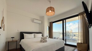 Quarto de hotel moderno com uma cama grande, uma pequena mesa de cabeceira, uma televisão de tela plana e uma grande janela com vista para a cidade.