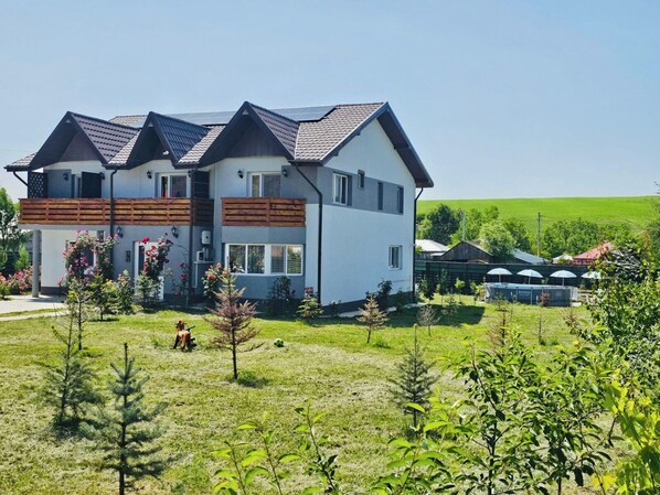 A two-story house with a sloped roof, a fenced yard, and a small tree in the foreground. - Cabana 15 persoane 7 camere  6 băi  Living pentru petreceri  Terase Grătar (Lețcani)