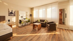 A modern hotel room with a bed, a sofa, a coffee table, a desk with a chair, and a large window. - ADAPT APARTMENT HOTEL Siegen (Siegen)