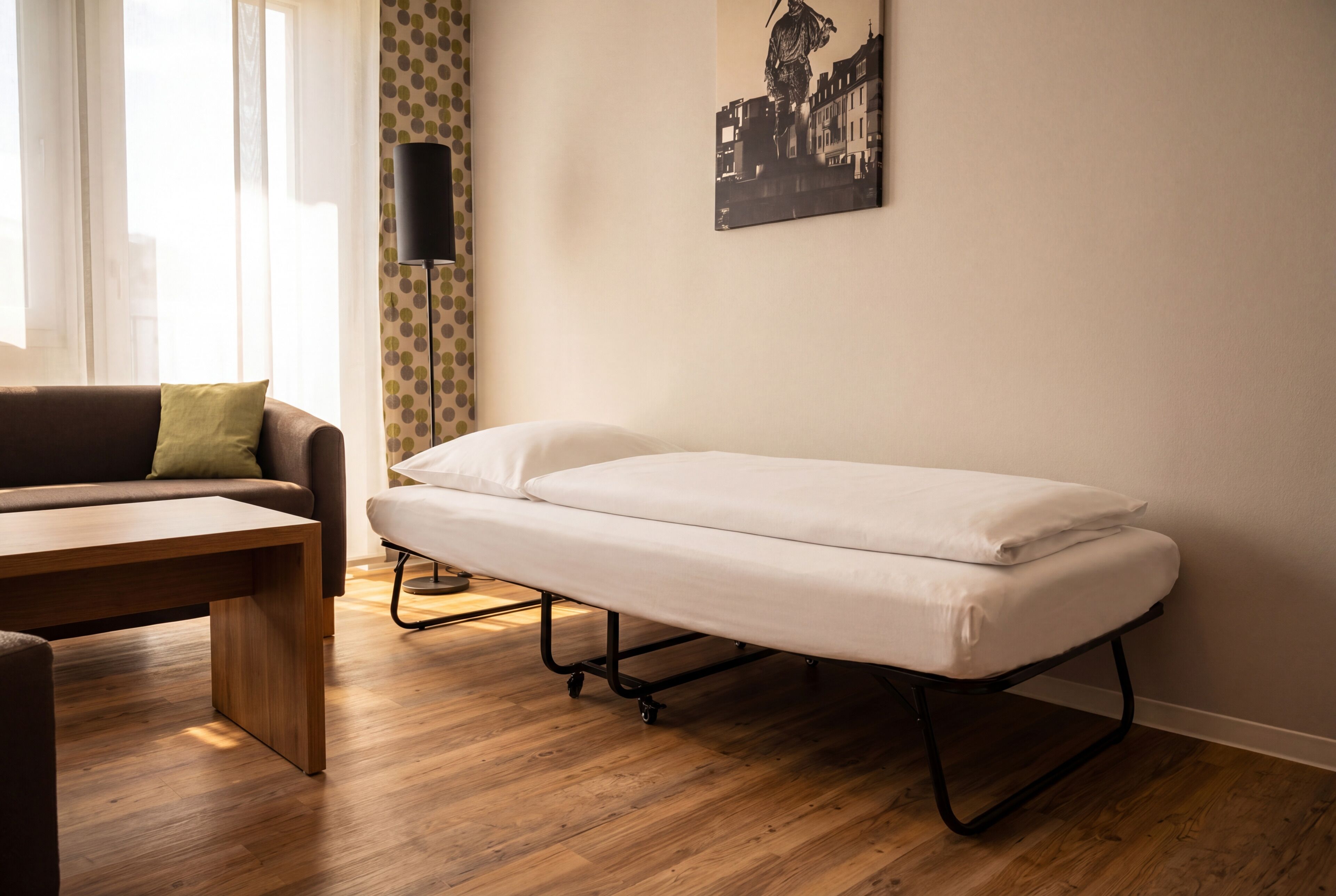 A room with a wooden floor, a sofa with a green pillow, a small table, a foldable bed, a wall-mounted lamp, and a cityscape painting.