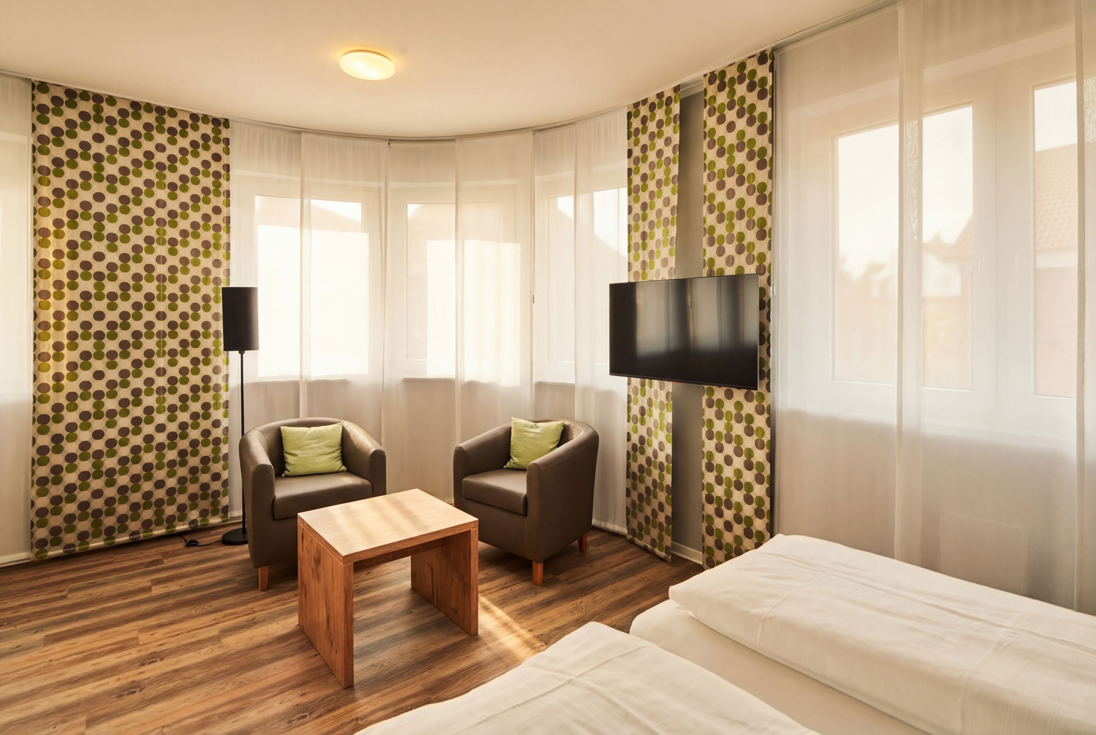 Habitación de hotel con suelo de madera, dos sillones, una mesita, una cama, un televisor y cortinas con estampado de lunares.