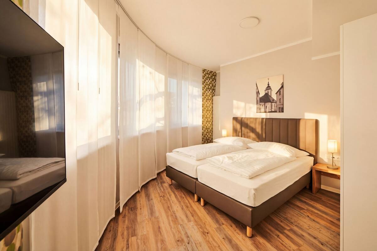 A hotel room with a bed, a bedside table with a lamp, a TV, and a window with curtains.