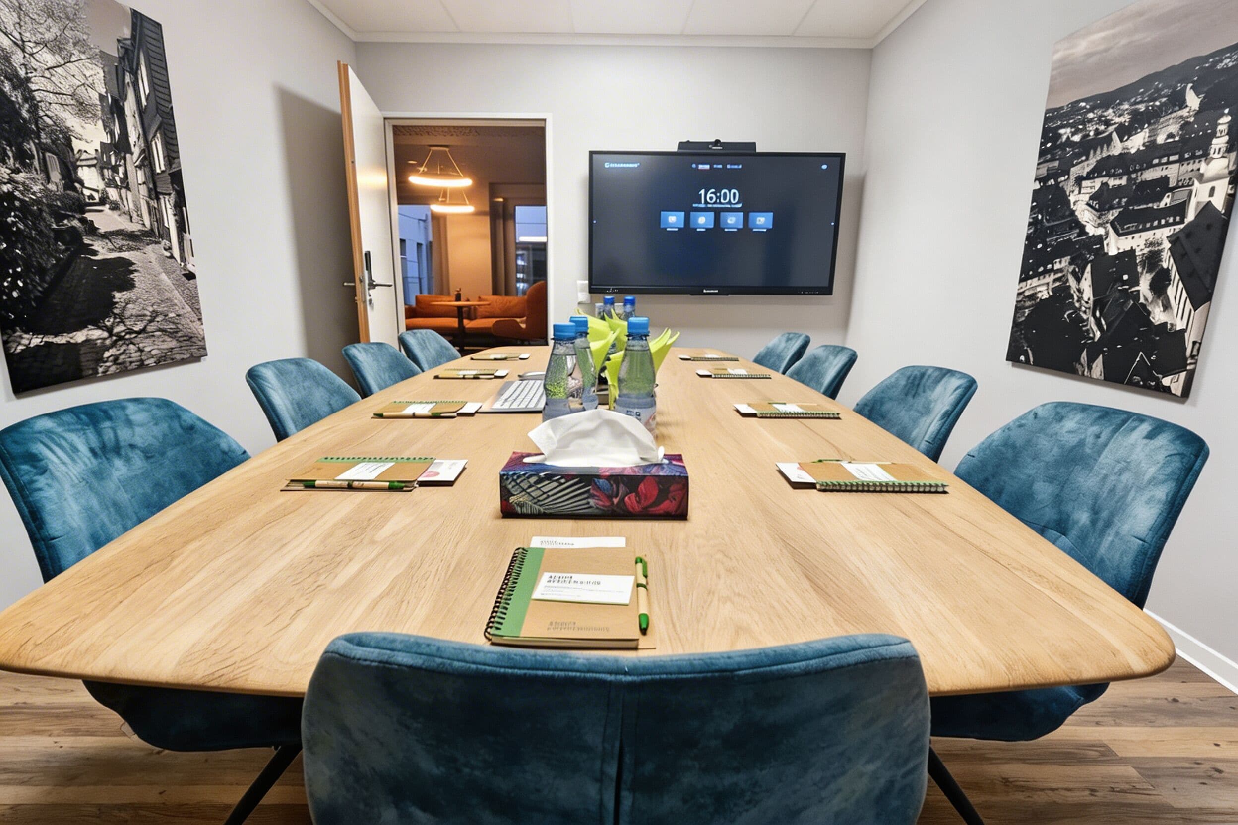 A conference room with a long table, blue chairs, a TV screen, and artwork on the walls.