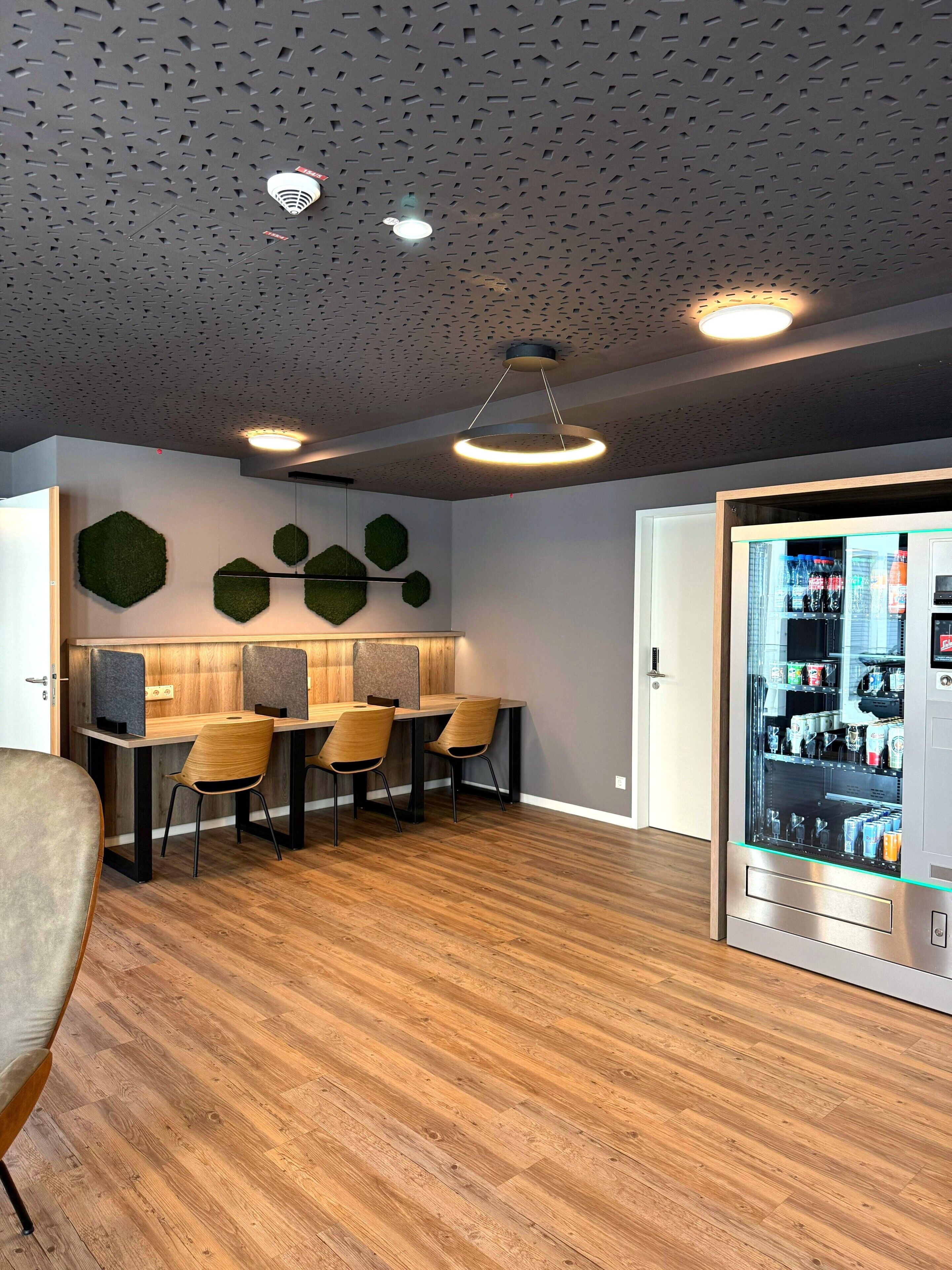 A modern interior with a wooden dining table, chairs, a vending machine, and a ceiling with a unique geometric pattern.