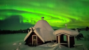 A snow-covered house with a thatched roof and a chimney, set against a backdrop of the Northern Lights. - Jokimaja Boutique Hotel (Kittila)
