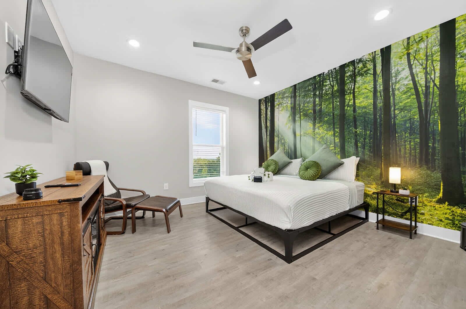 A bedroom with a bed, a desk, a chair, a ceiling fan, and a forest mural on the wall.