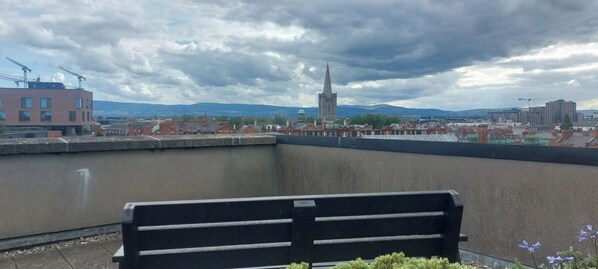 A balcony with a bench, overlooking a cityscape with buildings and a prominent tower.