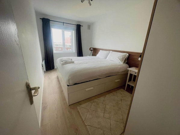 A bedroom with a bed, a bedside table, a window with curtains, and a view of buildings outside.