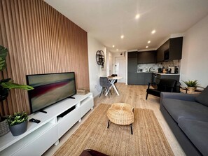 A modern living room with a flat-screen TV, a grey sofa, a woven ottoman, and a dining area with a table and chairs.