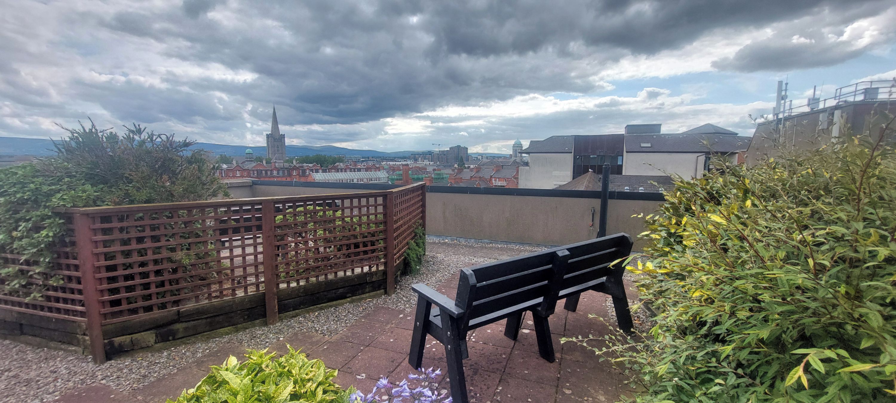 Une terrasse sur le toit, agrémentée d’une clôture en bois, d’un banc et offrant une vue sur le paysage urbain, notamment sur le clocher d’une église.