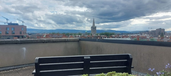 Eine Bank mit Blick auf eine Stadtlandschaft, darunter ein markanter Kirchturm, Baukräne und eine ferne Bergkette.