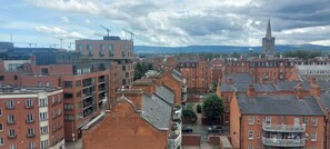 Une vue urbaine avec des bâtiments en briques rouges, le clocher d’une église et des grues de construction visibles au loin.