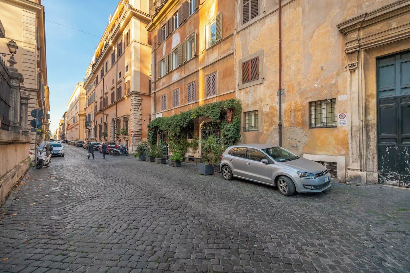 Une rue pavée bordée de bâtiments historiques, de voitures garées et de quelques piétons.