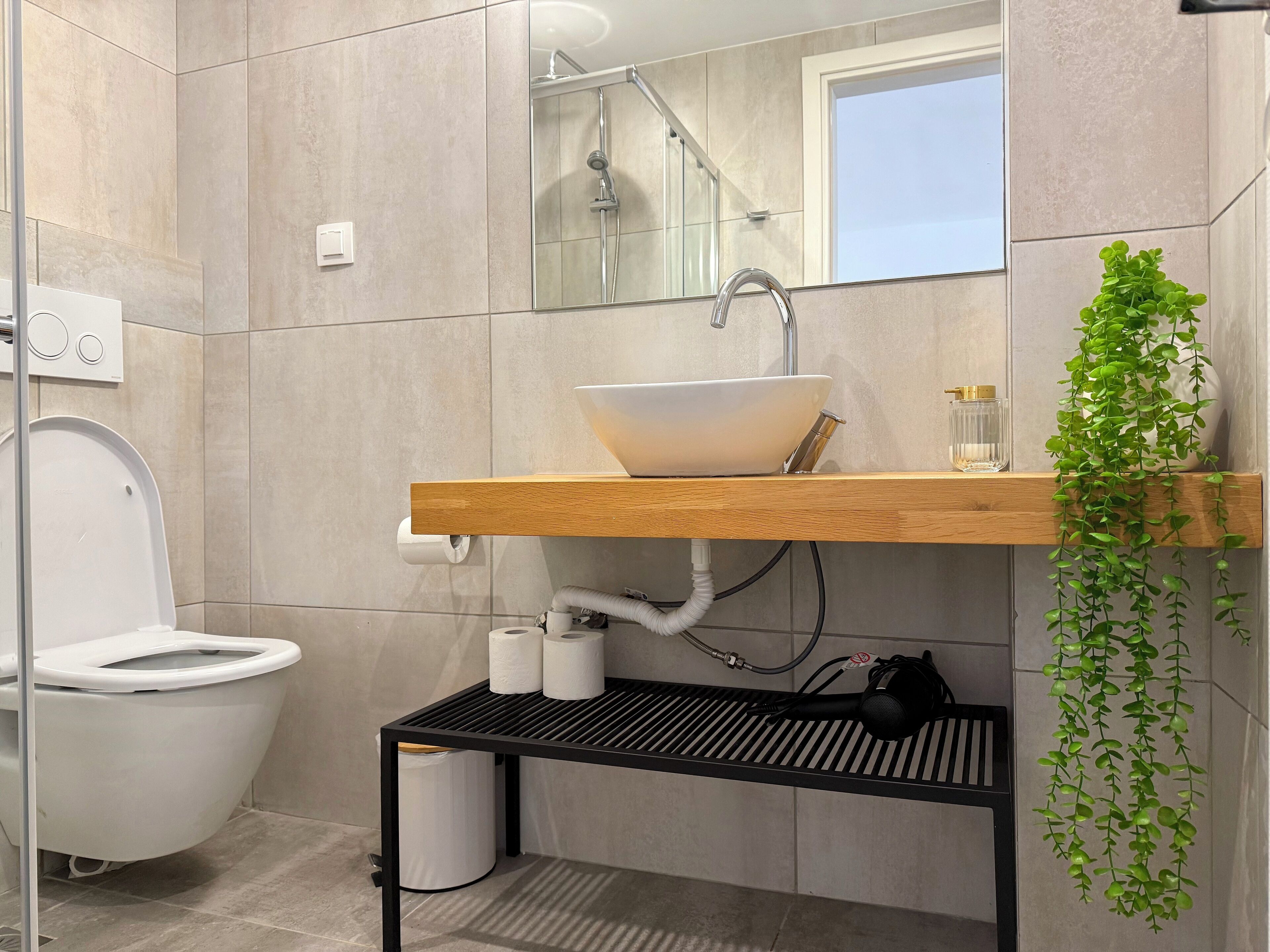 A modern bathroom with a white toilet, a sink with a wooden countertop, a mirror, and a green plant.