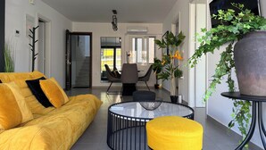 Una sala de estar moderna con un sofá amarillo, una mesita de centro redonda de cristal y un reposapiés amarillo. Hay un comedor con una mesa y sillas, y una planta grande en maceta.