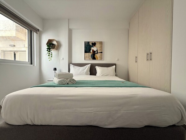 A neatly made bed with white linens, a green throw blanket, and rolled towels. There is a painting on the wall above the bed, a window on the left, and a wardrobe on the right. - TH0 - Tesla Smart Building - Cloud9 in the city (Athina)