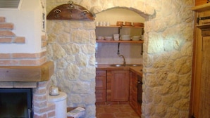 Un arco de piedra que da acceso a una cocina con chimenea, estantes con vajilla y armarios de madera.