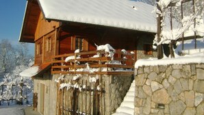 Una cabaña de madera con cimientos de piedra, un porche de madera y techo cubierto de nieve.