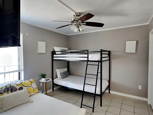 A bunk bed room with a ceiling fan, a small table with a plant, and a window with blinds.