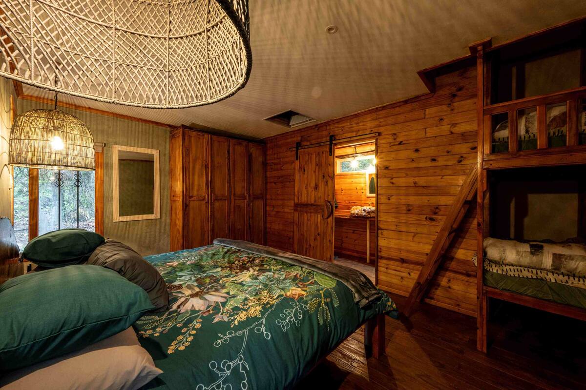A wooden cabin room with a bed, a hanging light fixture, and a window with a view of the forest.