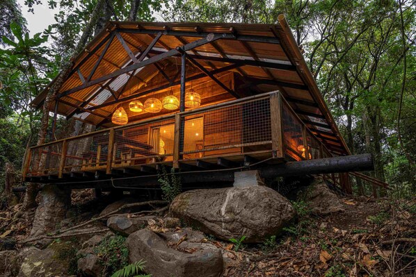 Eine rustikale Holzhütte mit Metallgeländer und hängenden Lichtern, umgeben von Felsen und Vegetation.