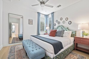 A bedroom with a large bed, a ceiling fan, a nightstand, a bedside table, and a mirror. - Vivid ATX Vibes | Pools. Personality. Pure Bliss! (Austin)
