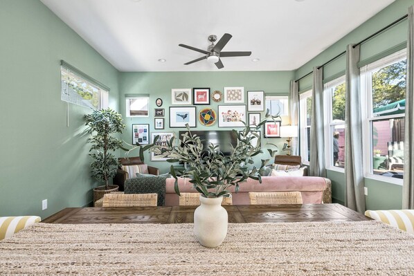 A living room with a ceiling fan, a large window, and a wall decorated with framed pictures. - Vivid ATX Vibes | Pools. Personality. Pure Bliss! (Austin)