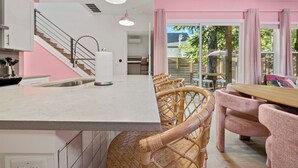 A modern kitchen with a white countertop, stainless steel sink, and a white vase. There are wicker chairs and a wooden table in the dining area. - Vivid ATX Vibes | Pools. Personality. Pure Bliss! (Austin)