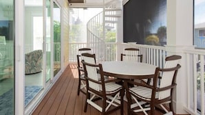 Une terrasse couverte avec une table ronde et des chaises, une porte vitrée donnant sur un jardin et une rambarde blanche.