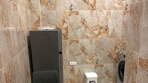 A compact room with a marble-tiled wall, a small table with a chair, a washing machine, and a compact refrigerator. - Luxury studio (Lomé)