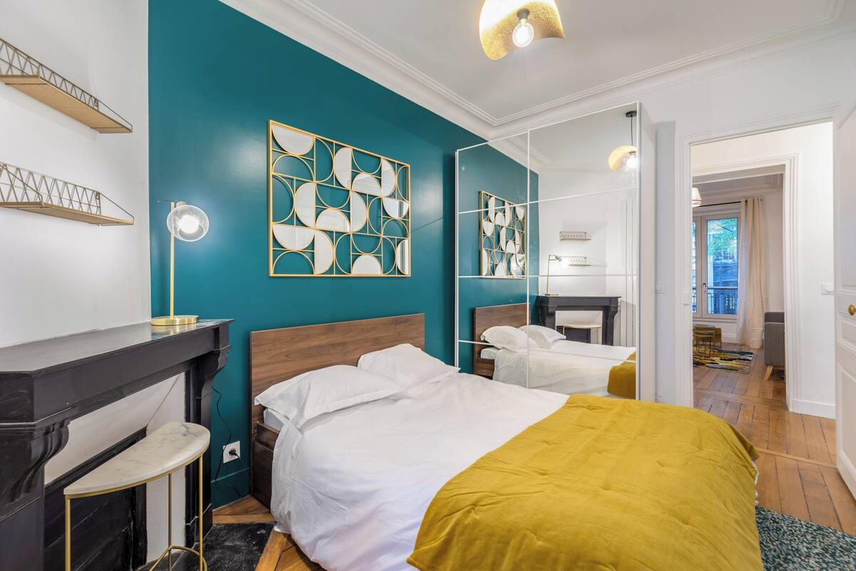 Une chambre moderne aux murs de couleur turquoise, avec un grand lit recouvert d’un plaid moutarde, un miroir et une petite table avec une lampe.