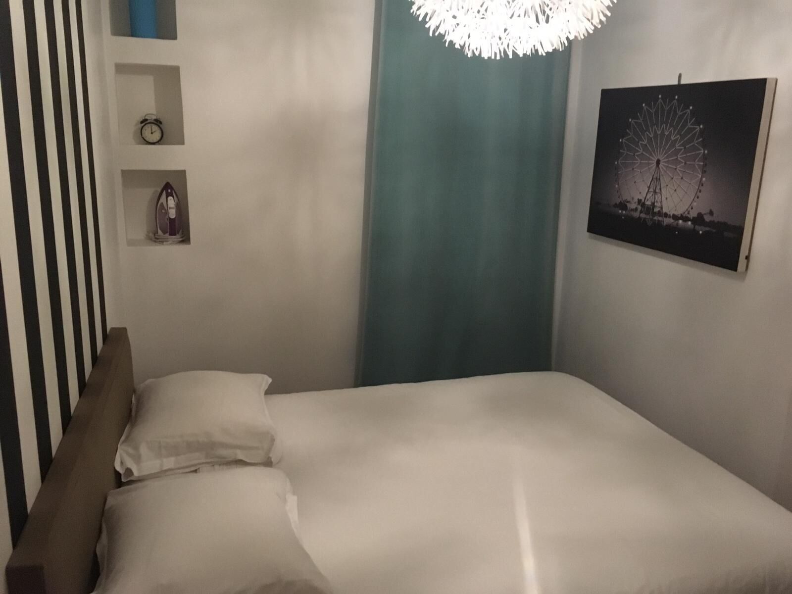 Un lit bien fait, avec une literie blanche, une tête de lit en bois, une horloge murale, une petite étagère avec une horloge et un objet décoratif, un rideau turquoise et un tableau encadré représentant une grande roue.