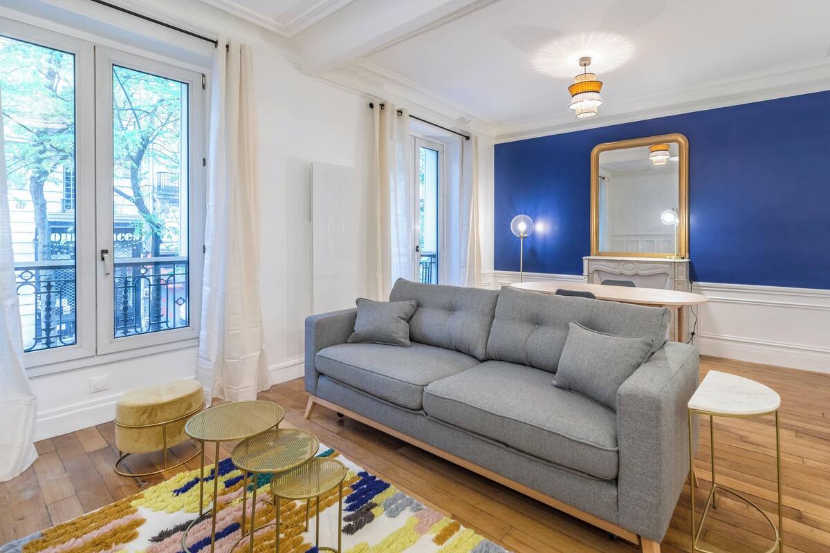Un salon moderne avec un canapé gris, une table basse ronde, un petit tabouret et un grand miroir sur un mur bleu.