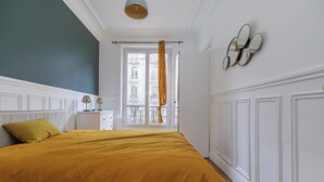 Une chambre avec un grand lit, une fenêtre avec des rideaux, une table de chevet avec une lampe et un miroir fixé au mur.
