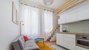 Une cuisine moderne avec des meubles blancs et un four en acier inoxydable, un canapé gris agrémenté d’un coussin rouge et une petite table ronde.