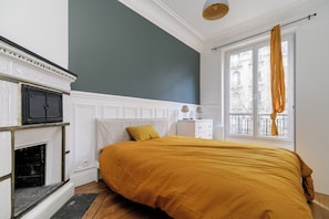 A bedroom with a bed, a fireplace, a window with curtains, and a view of buildings outside.