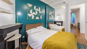 Une chambre moderne aux murs de couleur turquoise, avec un grand lit recouvert d’un plaid moutarde, un miroir et une petite table avec une lampe.