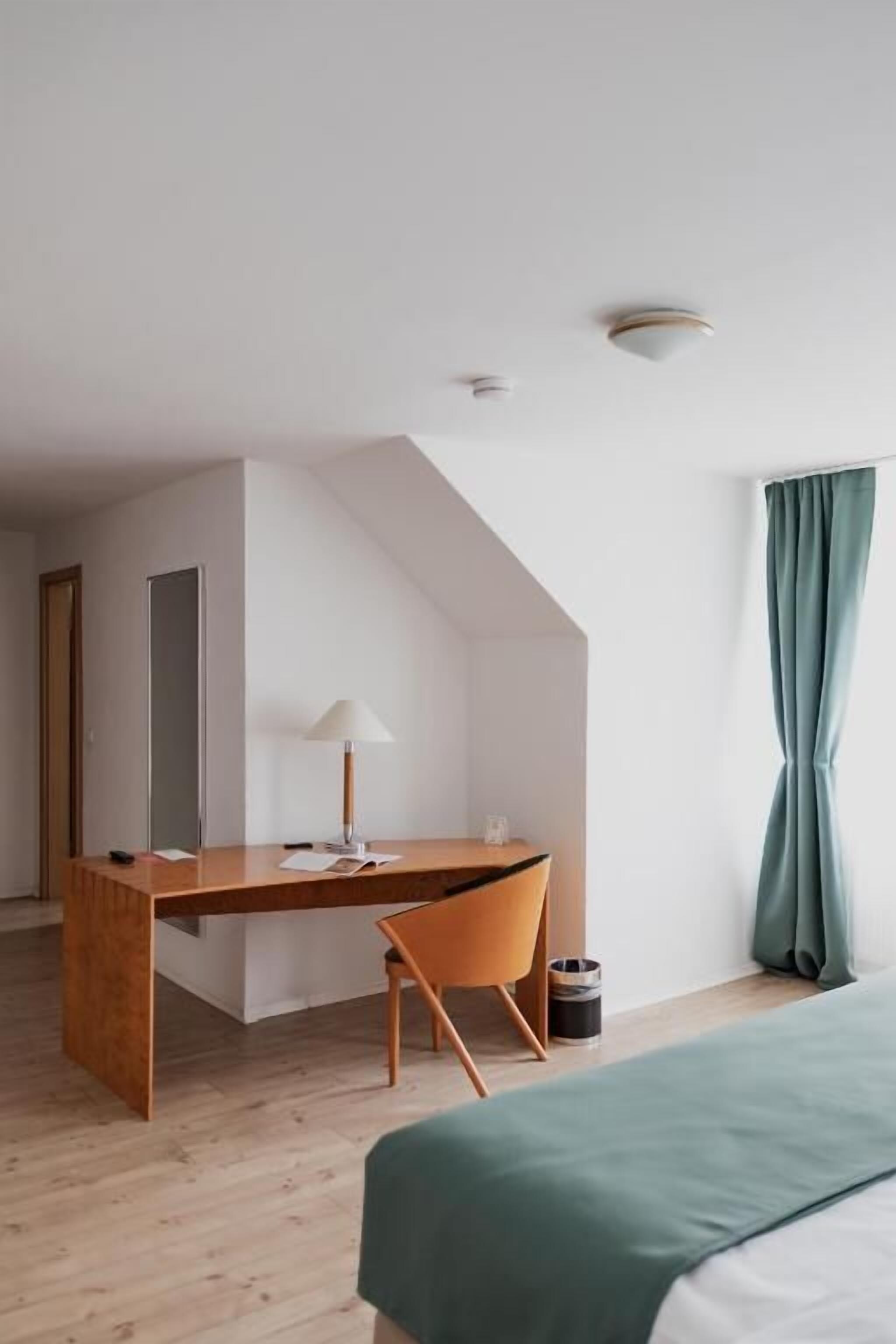 Una habitación de hotel moderna con un escritorio de madera, una silla, una cama con ropa de cama verde y un ventanal con cortinas.