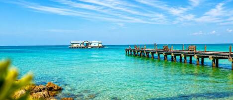 A clear blue sea with a wooden pier extending into it, a small overwater bungalow, and rocky foreground.