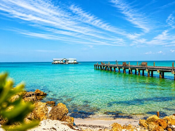 A clear blue sea with a wooden pier extending into it, a small overwater bungalow, and rocky foreground. - Shore to Please | Gulf Views 🌊 Elevator | Beach 1 Min 🌴 | Heated Pool & Spa 🌅 (Anna Maria)