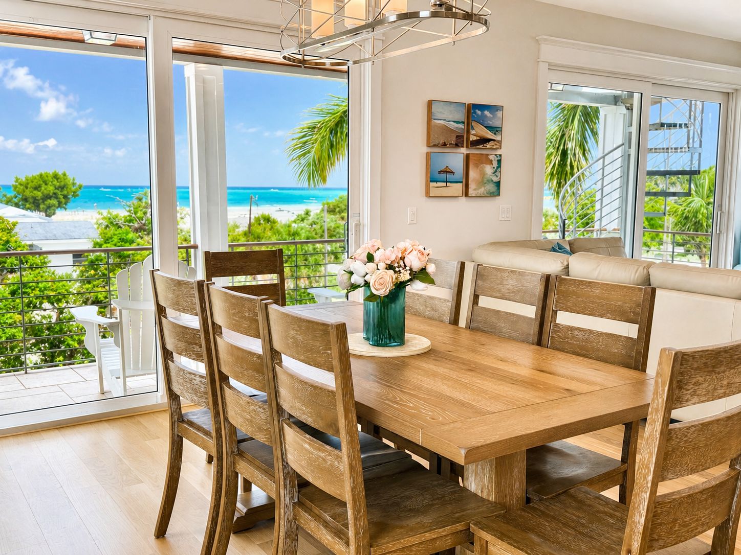 A dining area with a wooden table and chairs, a vase of flowers, and a view of the beach and ocean through large windows.